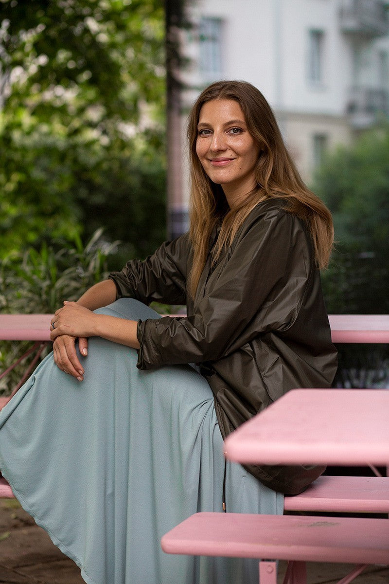 Kobieta w eleganckiej sukience i kurtce, siedząca na różowej ławce w zielonym ogrodzie – moda polska, self-care fashion